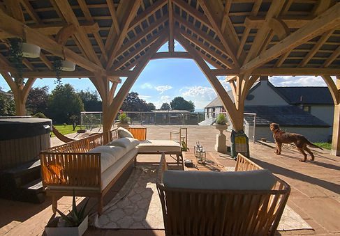 An oak pavilion with outdoor furniture and a hot tub, with a dog walking across the patio area.