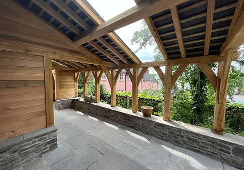 Covered stone patio with oak beam structure and a view of greenery outside.