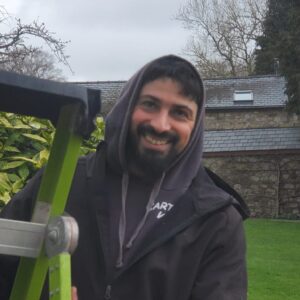 A smiling man with a beard, wearing a hooded jacket, standing outdoors near a green ladder by an oak tree.