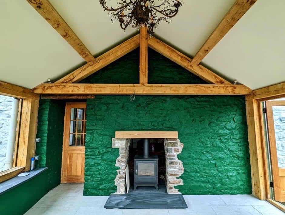 An a-frame room with a green textured wall and an oak wood-burning stove inset into a stone fireplace.