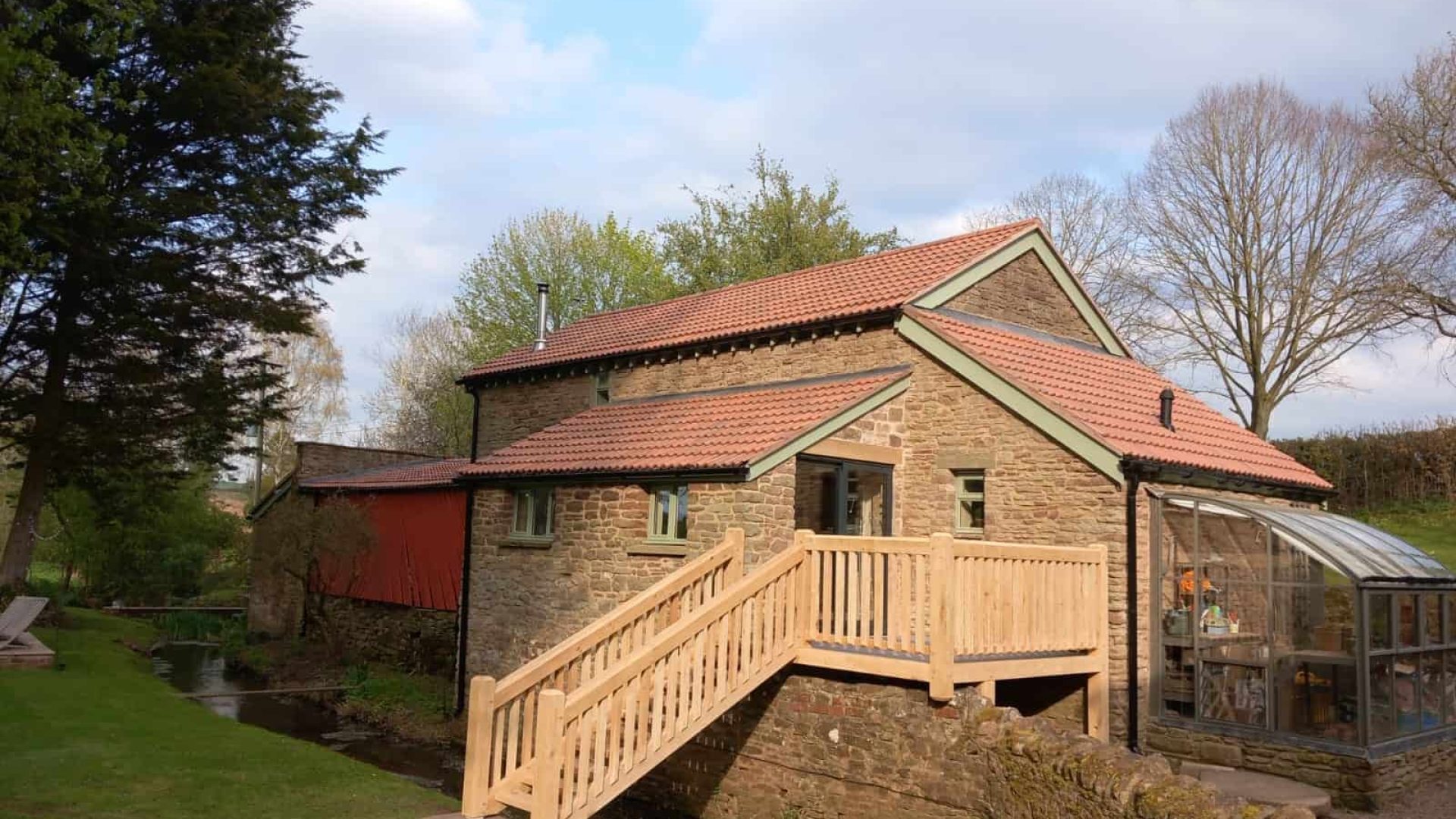 A stone cottage with a red tile roof featuring an oak deck and an attached greenhouse, set against a backdrop of greenery and a partly cloudy sky.