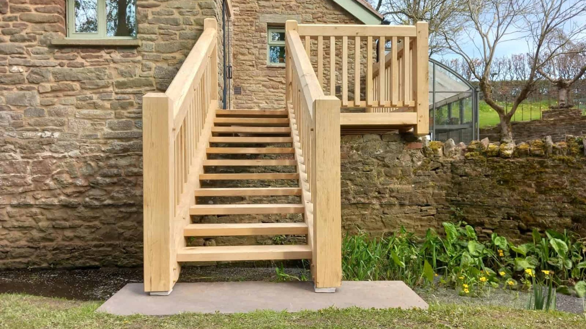 Wooden oak staircase leading to an elevated deck on the side of a stone building, surrounded by green grass.