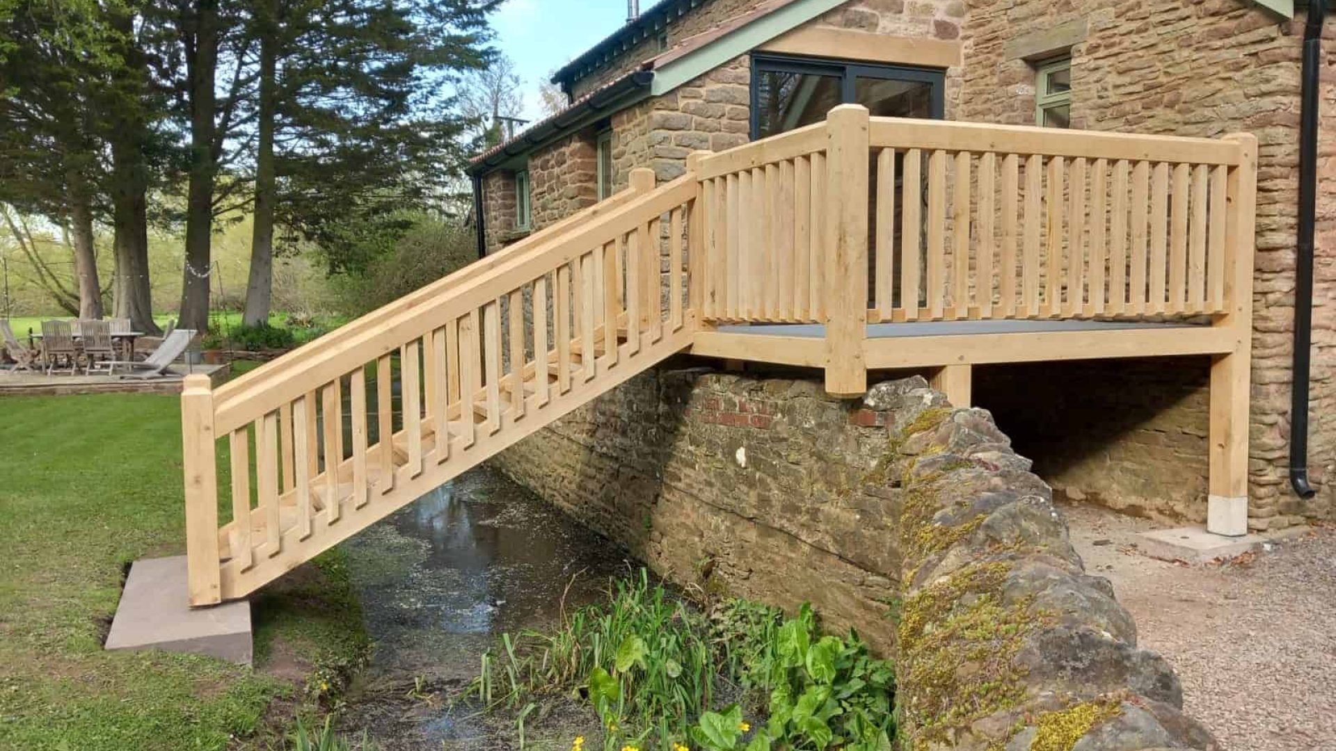 A wooden oak footbridge over a small stream next to a stone building.