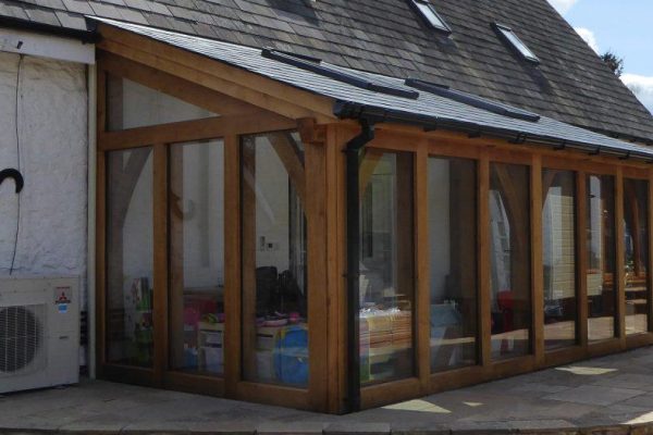 A modern sunroom extension with oak beams and glass panels attached to a stone building, featuring a heat pump unit on the side.